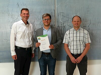 Drei Männer stehen nebeneinander vor einem abstrakten Kunstwerk. Der Mann in der Mitte hält ein Zertifikat in der Hand, während die anderen beiden ihm zugewandt sind. Sie tragen formelle Kleidung und stehen in einer hellen, modernen Umgebung.