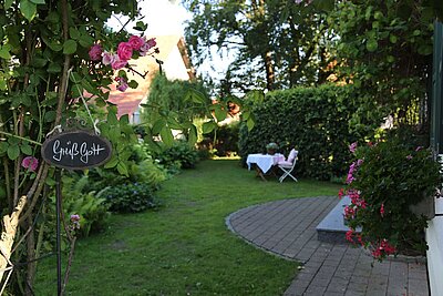 Ein schöner Garten mit grüner Rasenfläche und blühenden Pflanzen. Links hängt ein Schild mit der Aufschrift „Gott sei Dank“. Im Hintergrund steht ein Tisch mit weißen Tischdecken und zwei Stühlen, umgeben von Büschen und Bäumen.